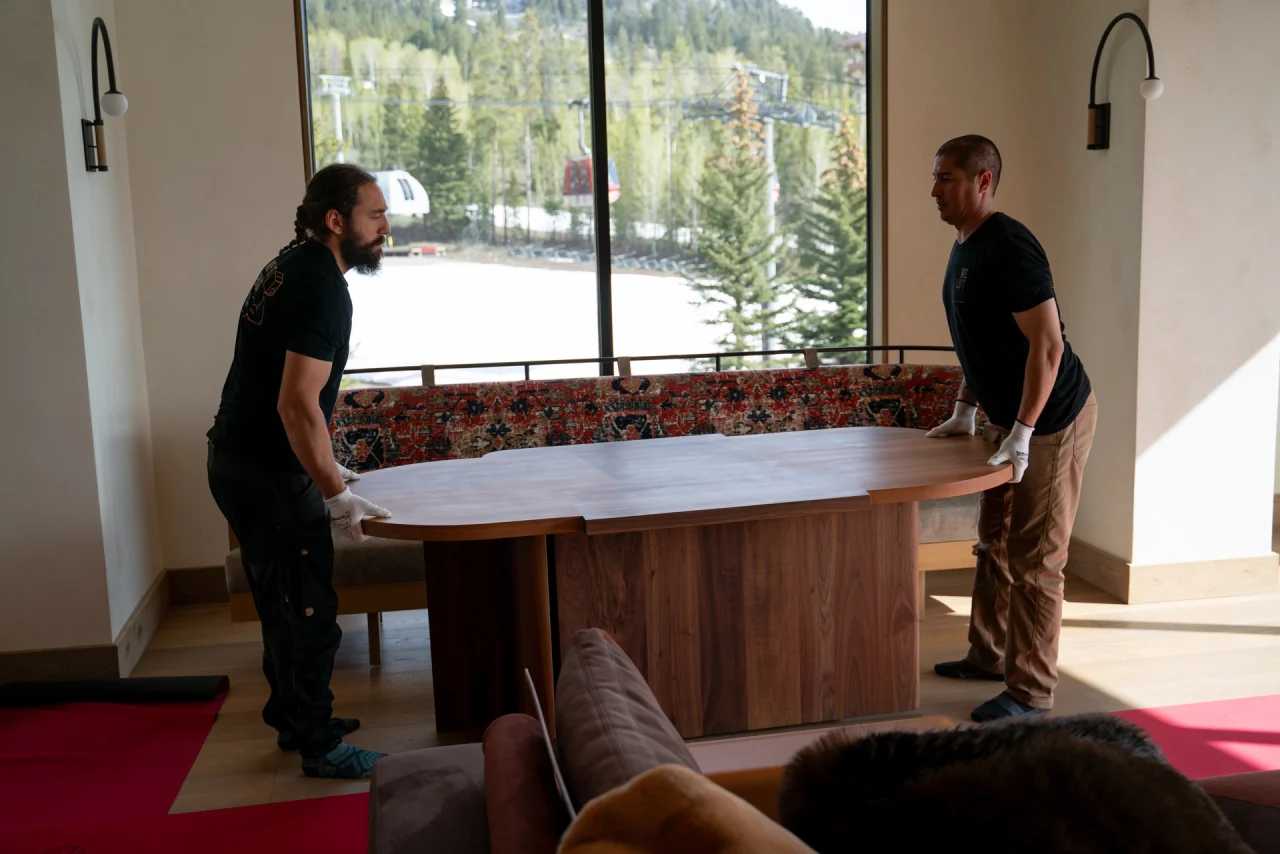 Two We Lift Things crew members in white gloves position a walnut dining table, with the Jackson Hole tram and gondola visible through the window behind them.
